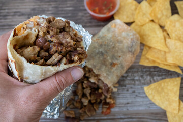 Hand holding a carne asada beef burrito wrapped in foil over a table with chips and salsa at a Mexican food restaurant. - obrazy, fototapety, plakaty