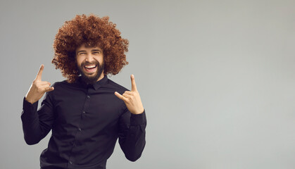 Yeah. Portrait of crazy cheerful businessman having fun on copy space background. Grey ad copyspace banner with funny handsome man with curly Afro hair doing horn sign and laughing. Rock party concept