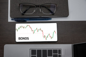 Buy or sell bonds concept. Top view of stocks price candlestick chart in phone on table near laptop, notepad and glasses with inscription bonds. Business, finance, commercial concept.