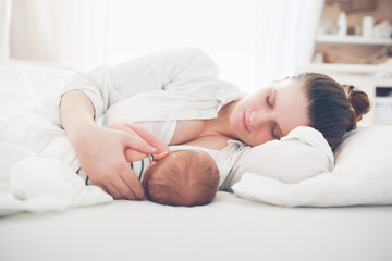 Obraz na płótnie Canvas Cute caucasian mom and newborn baby, mom breastfeeds her baby lying on a white background on the bed in the bedroom, close-up, soft focus