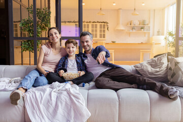 Smiling laughing parent showing funny video to son on television. Parent and son watching comedy movie on tv-set at home. Happy family sitting on comfort sofa. Cozy evening in living room interior