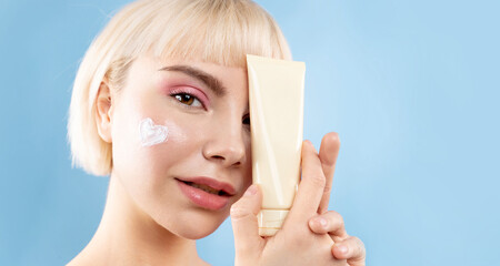 Smiling blond girl with shiny glowing skin, with cream on cheek,  beauty studio portrait on blue,...