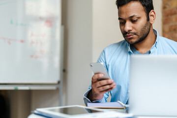 Fototapeta premium Middle eastern unshaven man using mobile phone while working with laptop