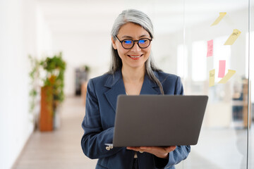 The smiling Asian woman in glasses holding a laptop in her hands while standing in a bright office