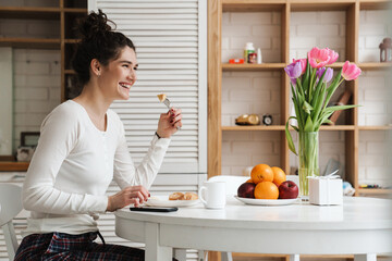 The side view of smiling woman bringing a piece of food while sitting at the table in the kitchen