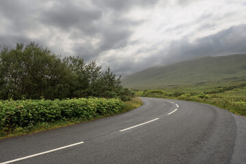 Small narrow asphalt road turn in Connemara. Beautiful mountain scenery in the background. Dramatic cloudy sky. Nobody. West of Ireland. Travel and transportation concept