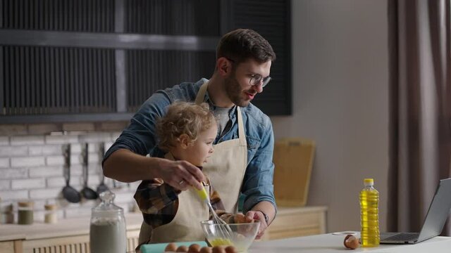 little son and father are viewing tutorial on laptop and cooking on home kitchen, whipping eggs in bowl, dad and child