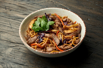 Delicious asian street food - egg noodles with beef, cilantro, vegetables and scrambled eggs in a white bowl on a wooden background. Wok noodles