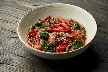 Mouth-watering Asian street food - beef in hot soy sauce with broccoli, chili, and sesame seeds in a bowl. Wood background