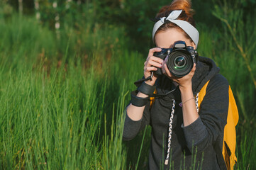 A girl in a gray jacket with a yellow backpack holds a professional photo-video camera in her hands. Against the background of green nature and forest.