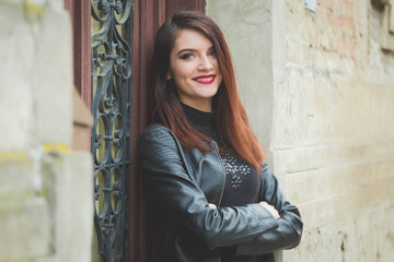 Beautiful Caucasian female with red lipstick and black leather coat posing near long wooden door