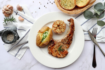 Grilled pork chop steak with baked potato, roasted garlic in white plate and garlic bread on the side - Top view on marble background
