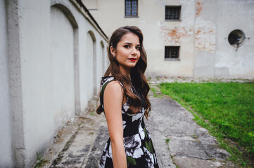 Beautiful brunette in dress goes in the courtyard of the old castle. Stylish girl in black dress on background of gray vintage building. Big sad eyes.