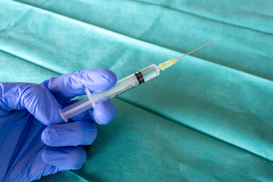 Health Care worker with blue surgical gloves holding a syringe in front of a green surgical fabric. Surgeon hand with a vaccine. A syringe with the vaccine. 