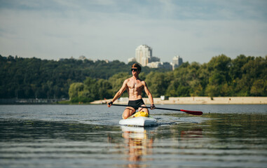 Handsome athletic man with muscular body swims on sup board on pond on summer hot day, wears...