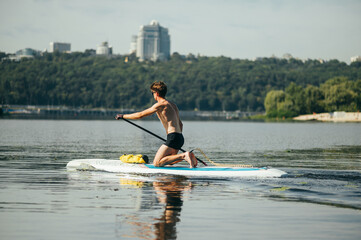 Athletic young man surfing on a sup board on the river in the summer on a weekend, actively rowing, rear view.