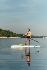 Handsome young man with a naked torso stands on a sup board with an oar in his hand and floats on the river, looking to the side. Active recreation on the water.