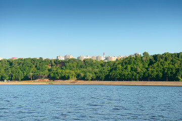 Summer landscape with a river bank with a concrete bank, green forest and high-rise buildings