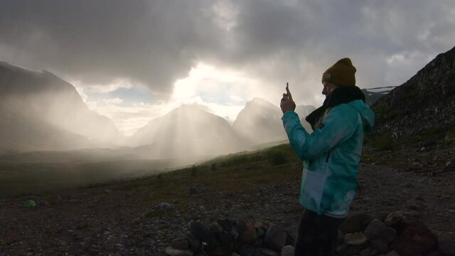 Female hiker taking photos with her phone of the big mountain Kebnekaise at sunset. Kebnekaise fjällstation.