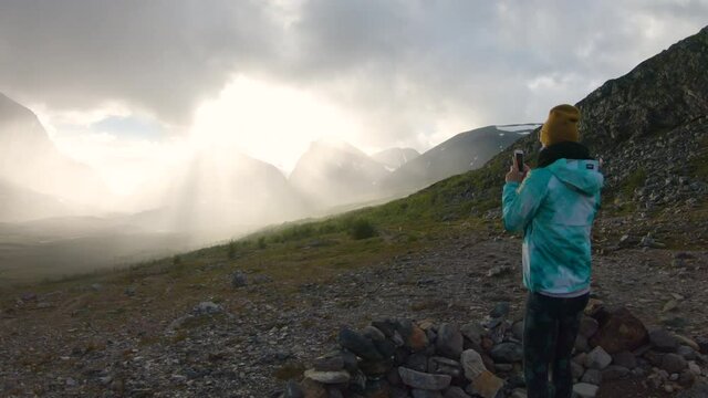 Female hiker standing alone and taking photos with her phone of the big mountain Kebnekaise at sunset. At Kebnekaise fjällstation Northern Sweden.