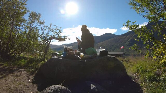 Female hiker eating breakfast on a big rock in the nature. You can see mountains and a helicopter flying by in the background.