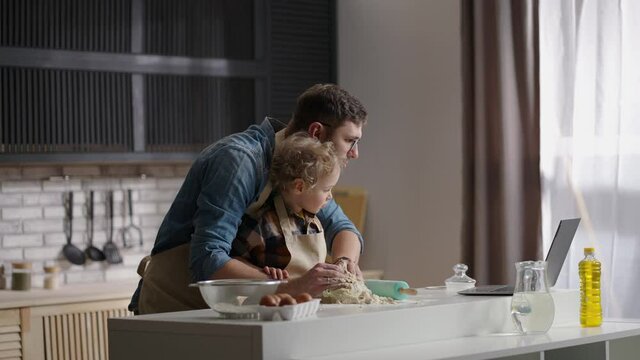 single father is learning his little son to cook, reading recipe in internet by laptop, cooking dough at home kitchen