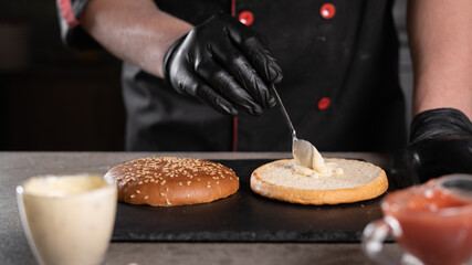 step by step recipe. Stage 2. The chef prepares a delicious burger. Close-up of hands in black gloves putting white sauce on a bun, menu, delicious and unhealthy food.