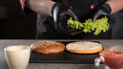 step by step recipe. Stage 3. The chef prepares a delicious burger. Close-up of a hand in black gloves putting a green leaf of lettuce on a bun with sauce, menu, delicious and unhealthy food.