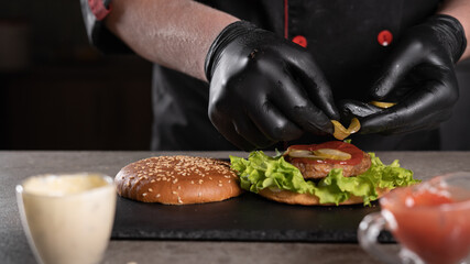 step by step recipe. Stage 6. The chef prepares a delicious burger. Close-up of a hand in black gloves putting a pickled cucumber on a cutlet, menu, delicious and unhealthy food.
