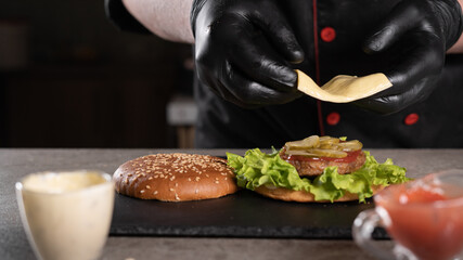 step by step recipe. Stage 7. The chef prepares a delicious burger. Close-up of a hand in black gloves putting a piece of cheese on a pickled cucumber, menu, delicious and unhealthy food.
