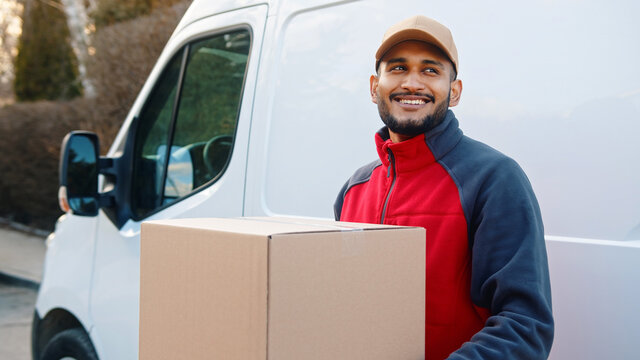 Delivery man smiling while holding parcel in front of transportation van. High quality photo