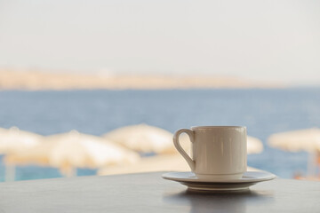 White cup of coffee on the table overlooking the beach with white umbrellas and the blue sea.