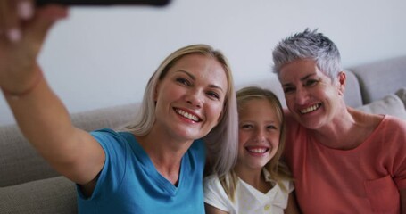 Caucasian grandmother, mother and daughter smiling while taking a selfie with smartphone at home - Powered by Adobe