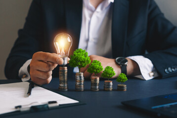 Businessman holding a light bulb and stacks of money growing with green trees. Concept of
pass and increase of renewable energy. Alternative sources of energy. Green energy, eco energy. 