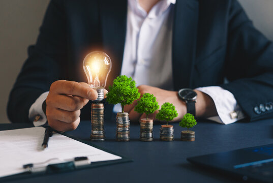 Businessman Holding A Light Bulb And Stacks Of Money Growing With Green Trees. Concept Of
Pass And Increase Of Renewable Energy. Alternative Sources Of Energy. Green Energy, Eco Energy. 