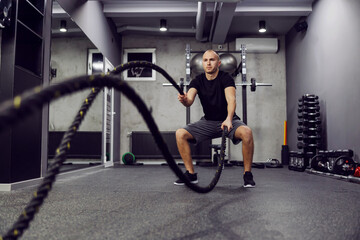 Fototapeta na wymiar Man training with battle rope in cross fit gym. Strong muscular man working out with battle ropes. Side view photo of fitness man in fashionable sportswear. Strength and motivation