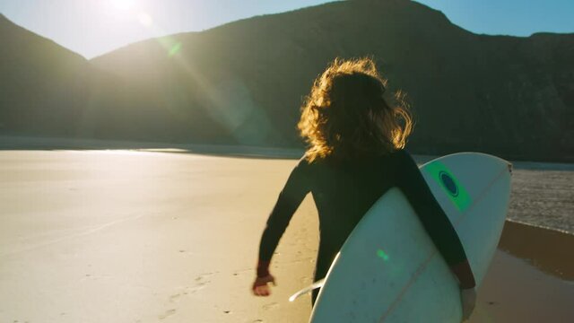 Slow motion shot of beautiful young woman running with surfboard on secluded epic beach. Real and candid portrait of authentic female surfer at adventure or surfing camp