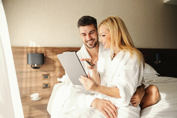 Romantic couple weekend. Sunny morning in a hotel room, a man and a woman making an online reservation in the comfort of a bathrobe. A woman holding a payment card and a tablet in the hugs of men