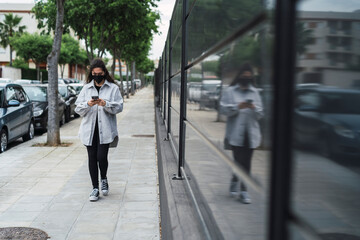 Chica joven con mascarilla negra por covid 19 usando el smartphone en la calle junto a una pared negra