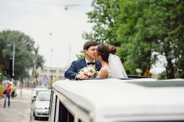 Gorgeous elegant wedding couple kissing and hugging on the roof of a white limousine on a city...