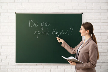 Teacher near green chalkboard with inscription Do You Speak English? in classroom