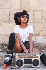 Portrait of a black woman with afro hair, roller skates and sunglasses listening to music with headphones from an old radio cassette player while sitting on a stone stairs outdoors on a hot summer day