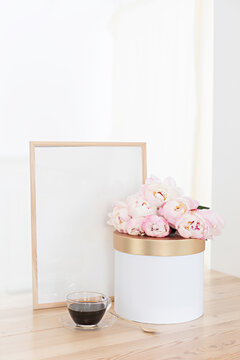 Vertical frame and gift box mockup on a wooden table in the kitchen. Glass jug with a bouquet of pink peonies and a cup of black coffee. Scandinavian style interior.
