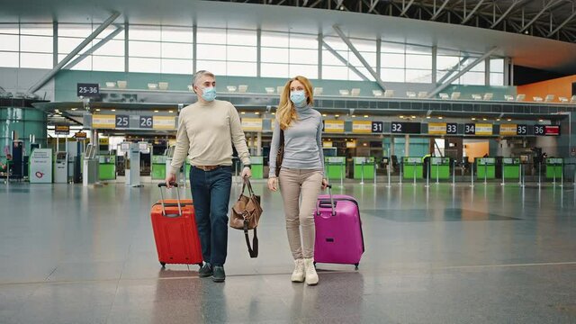 Couple of confident middle aged travellers wearing protective face mask walking with bright suitcases through airport
