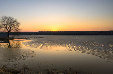 Golden sunset on a lake with tree silhouette and small pier in the foreground. Clear sky and trees in the background. Remains of half-melted ice form fancy pattern on the water.