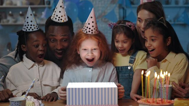 Crop View Of Excited Little Kid With Red Hair Opening Present Box And Smiling. Diverse Cheerful Children, Man And Woman In Festive Hats Standing Near Birthday Girl While Having Party.