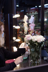 Florist works with colors. Flower seller is holding an empty sign for the price of roses. Flowers shop worker in a mask standing in flower shop and checking flowers in glass vase.