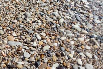 Top view of round flat colorful pebbles, sand and seashells, wet by sea waves. Natural marine pattern, half part of the frame blurred under water. Fresh summer holiday inspiration, relaxation mood.