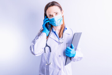 Caucasian young woman with stethoscope gown and gloves and mask holding mobile phone to one ear and reports in the other. technology and medicine communication concept