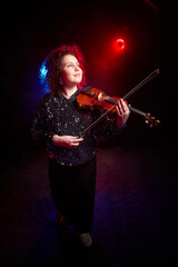 Fototapeta premium A brunette curly girl in black shiny clothes and with violin on a black background. Model, actress and violinist posing in the studio